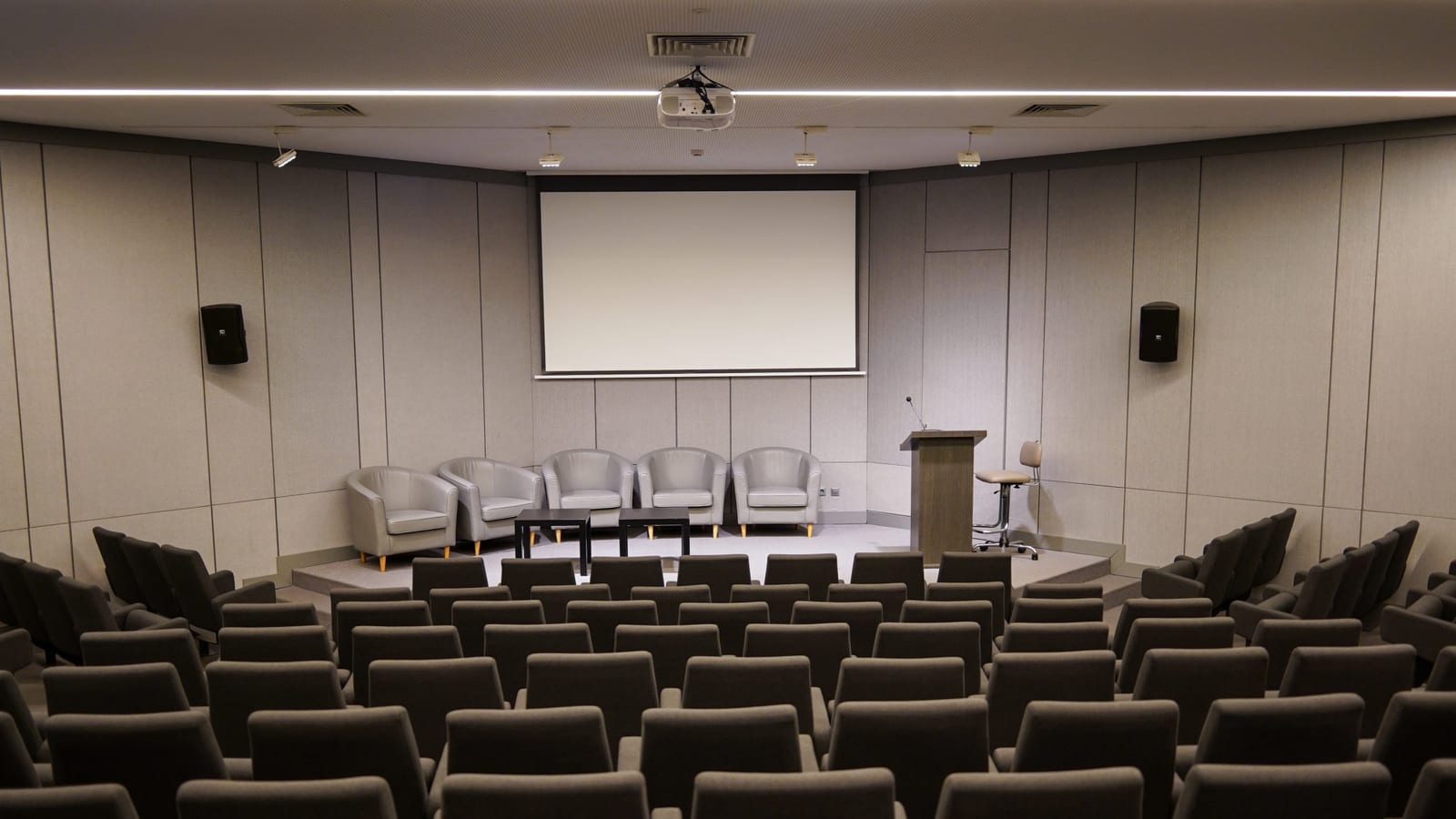 Photograph of an empty conference room with rows of chairs facing a stage area. there are five gray chairs and two small